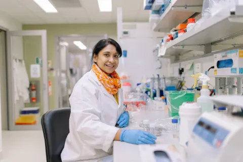 Women in a lab coat sitting at a desk in a lab smiling at the camera. 
